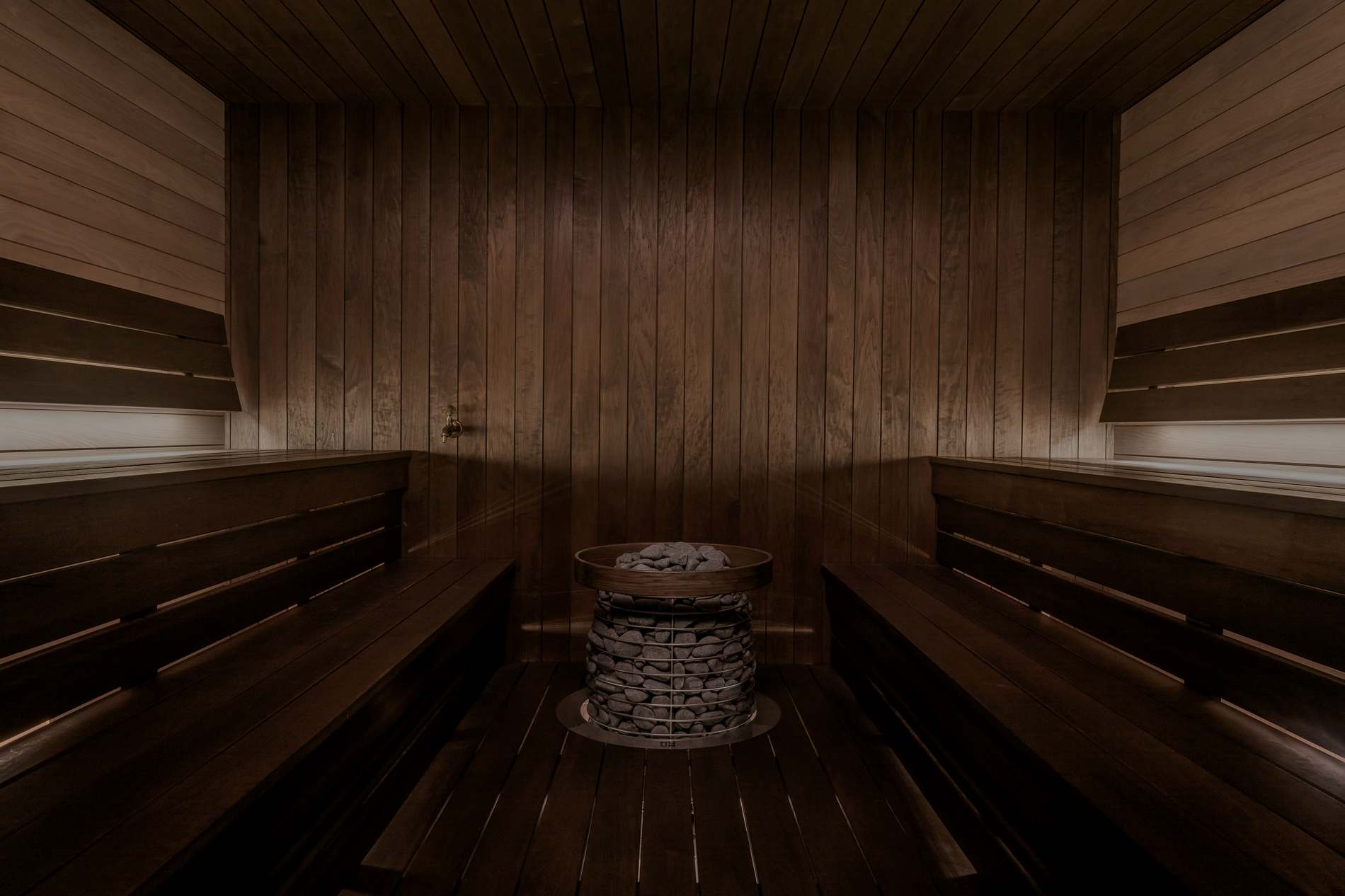 Dark cedar sauna interior with a stone-topped heater and dual benches, dim ambient lighting